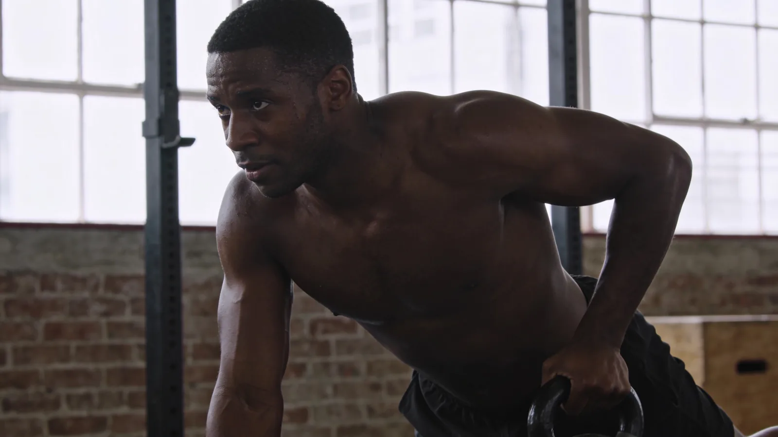 Shirtless muscular man bent forward gripping a kettlebell in a brick-walled gym with large windows.