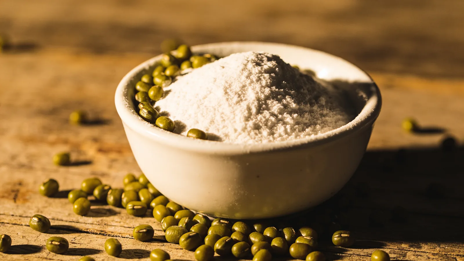 White bowl of pea protein powder surrounded by scattered green legumes on a rustic wooden table