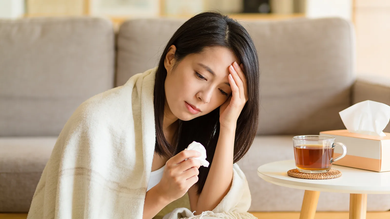 Woman wrapped in a blanket holding her head, looking nauseous beside tea and tissues on a table