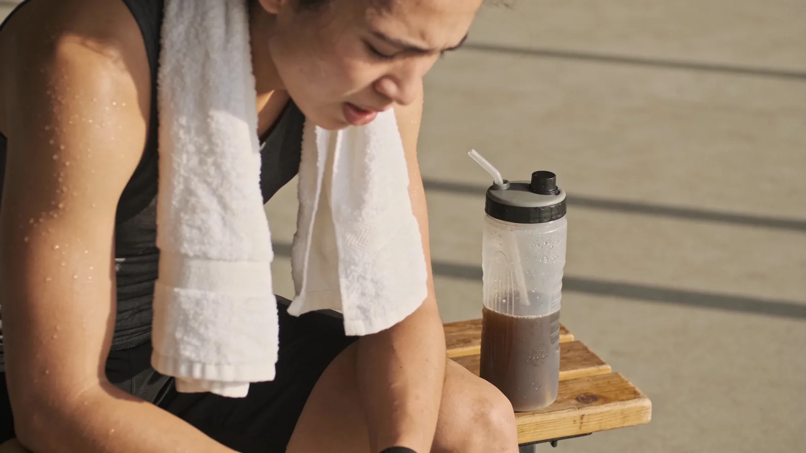 Sweating athlete resting with a towel beside a shaker bottle of brown protein shake after training
